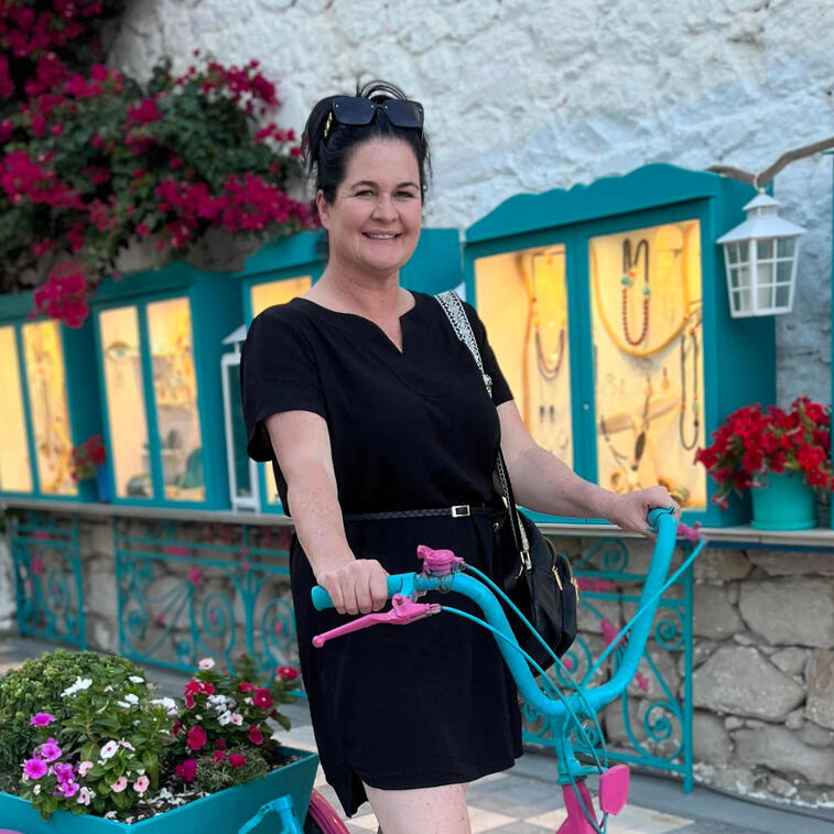 Lady with a black dress holding a pink and blue bicycle in front of shop windows with jewelry inside. Pink Bougainvillea flowers, red flowers and white flowers in the background in Milos Greece, photo taken by Mia's Photography and Events.