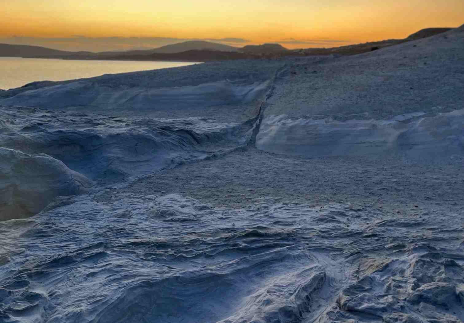 Sarakiniko sunrise in Milos Greece, photo taken by Mia's Photography and Events. The sun is starting to rise on the horizon with shades of orange in the sky, image also contains white volcanic rock landscape.