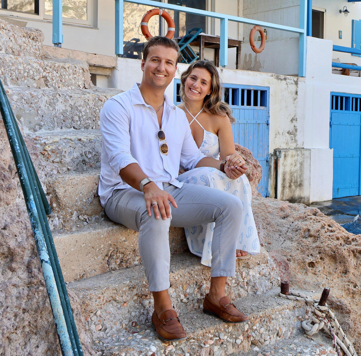 Photo of couple posing for a photo shoot in Milos Greece, photo taken by Mia's Photography and Events. The landscape background of this photo is Firopotamos beach with a church and small fishing village.