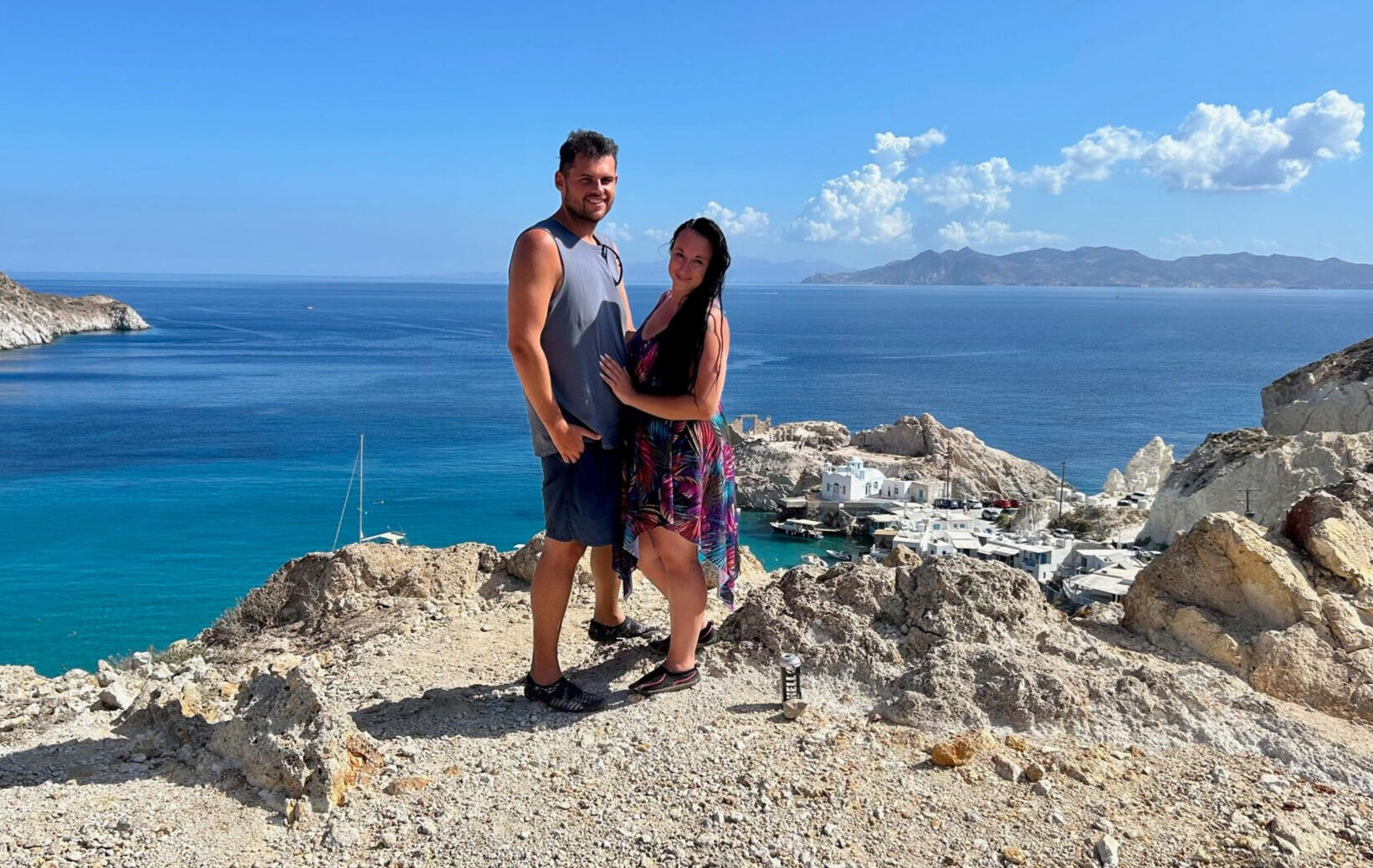 Photo of couple posing for a photo shoot in Milos Greece, photo taken by Mia's Photography and Events. The landscape background of this photo is Firopotamos beach with a church and small fishing village.