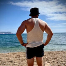Solo destination photo shoot of man standing on beach, wearing sunglasses, a black hat, white shirt and black shorts with sea and stones in the background in Milos Greece, photo taken by Mia's Photography and Events.