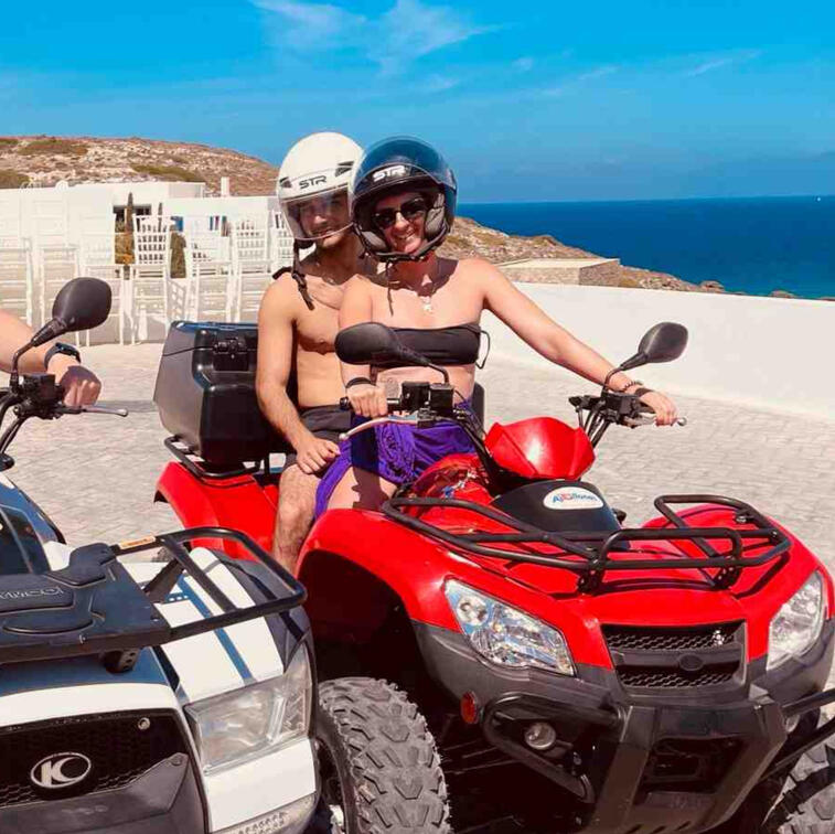 2 people on ATV in Milos Greece, one woman and one man, photo taken by Mia's Photography and Events. One white ATV, one red ATV. Smiling for a photo shoot. Blue sea in the background.
